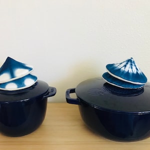 Puede incluir: Dos ollas de hierro fundido esmaltado azul con tapas. Las tapas están coronadas con círculos de tela azul y blanca, que se asemejan a una pila de origami.
