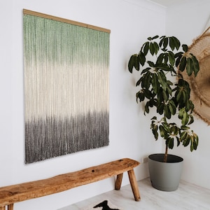 Puede incluir: Un tapiz de macramé de color verde, blanco y gris degradado con una barra de madera en la parte superior. El tapiz está colgado en una pared blanca junto a un banco de madera y una planta en maceta.