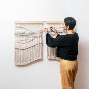 Puede incluir: Dos tapices de pared tejidos con una paleta de colores neutros y naturales. Los textiles presentan diversas texturas y detalles de flecos, montados sobre varillas de madera. Se muestra a una persona ajustando uno de los tapices, lo que sugiere una decoración del hogar y arte textil.