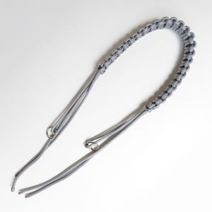 Puede incluir: Una correa de cámara de paracord gris con un asa trenzada y dos cordones ajustables con anillos metálicos para la fijación. La correa está diseñada para ser duradera y cómoda.