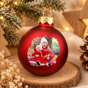 Pode incluir: Um enfeite de Natal esférico vermelho mate com uma impressão fotográfica personalizada na frente. A imagem mostra duas crianças, uma com um fato de neve vermelho e outra com um casaco cor-de-rosa, sentadas ao ar livre. Tem uma tampa dourada.