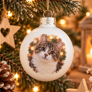 Pode incluir: Um enfeite de Natal redondo e branco com uma imagem impressa de um gato malhado castanho e branco na parte frontal. O enfeite tem uma tampa de metal prateado com gravações de flocos de neve e está suspenso por um fio dourado.