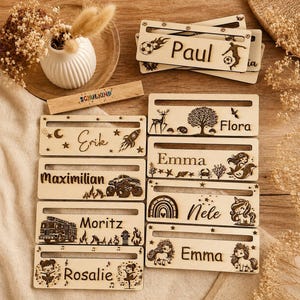 Könnte beinhalten: Personalisierte Namensschilder aus Holz mit den Namen "Paul", "Erik", "Maximilian", "Moritz", "Emma", "Flora", "Nele" und "Rosalie". Die Schilder sind mit Motiven wie Raketen, Feuerwehrautos und Einhörnern verziert und aus hellem Holz gefertigt.