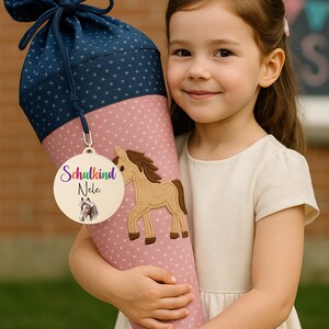 Pode incluir: Uma grande mochila escolar em forma de cone em rosa e azul marinho, decorada com um aplique de cavalo e uma etiqueta de madeira que diz "Schulkind Nele". A mochila &eacute; segurada por uma menina.