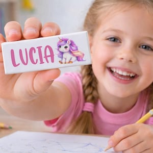 May include: A white rectangular name tag with the name "LUCIA" in pink script and a cartoon unicorn illustration. The unicorn has a purple mane and tail. A young girl is holding the name tag and smiling.