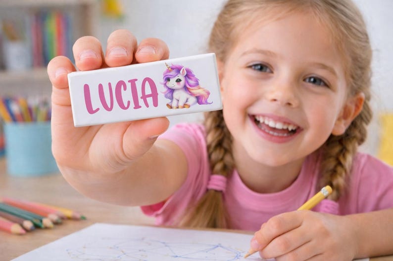 May include: A white rectangular name tag with the name "LUCIA" in pink script and a cartoon unicorn illustration. The unicorn has a purple mane and tail. A young girl is holding the name tag and smiling.