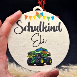 Pode incluir: Enfeite redondo de madeira com as palavras "Schulkind Eli" e uma ilustra&ccedil;&atilde;o de um monster truck. O enfeite tem uma pequena faixa de bandeiras coloridas e um furo para pendurar. Di&acirc;metro: aprox. 8 cm.