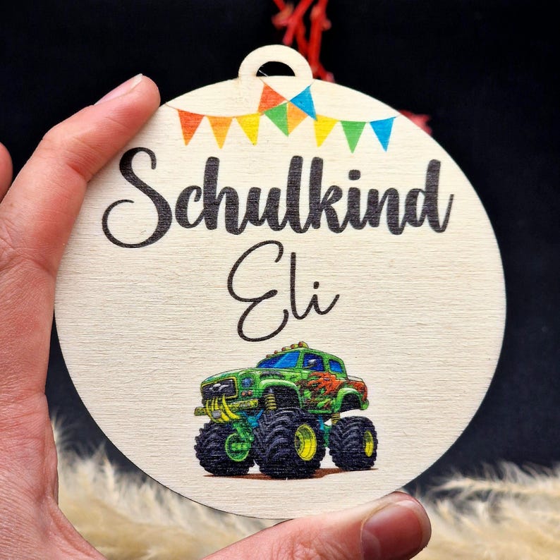 Pode incluir: Enfeite redondo de madeira com as palavras "Schulkind Eli" e uma ilustra&ccedil;&atilde;o de um monster truck. O enfeite tem uma pequena faixa de bandeiras coloridas e um furo para pendurar. Di&acirc;metro: aprox. 8 cm.