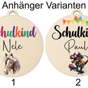 Pode incluir: Dois ornamentos redondos de madeira com um furo no topo. Um apresenta o texto "Schulkind Nele" em cores do arco-&iacute;ris, uma ilustra&ccedil;&atilde;o de cavalo e o n&uacute;mero 1. O outro tem "Schulkind Paul", um dinossauro num skate e o n&uacute;mero 2.