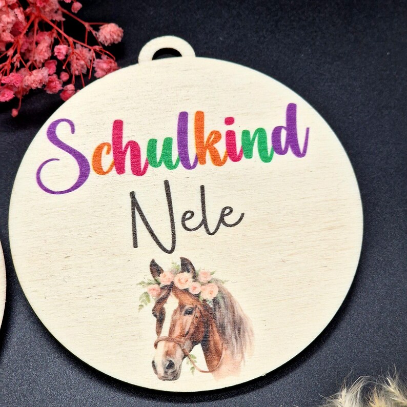 Pode incluir: Enfeite redondo de madeira com furo para pendurar. Apresenta a palavra "Schulkind" em cores do arco-&iacute;ris, o nome "Nele" em preto e uma ilustra&ccedil;&atilde;o em aquarela de um cavalo adornado com uma coroa de flores.