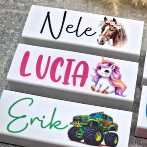 May include: Three white rectangular blocks with names and illustrations. The top block says "Nele" with a horse wearing a floral crown. The middle block says "Lucia" with a unicorn. The bottom block says "Erik" with a monster truck.