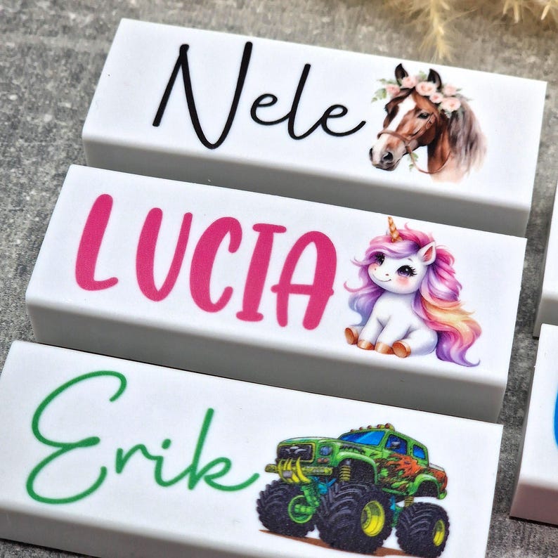 May include: Three white rectangular blocks with names and illustrations. The top block says "Nele" with a horse wearing a floral crown. The middle block says "Lucia" with a unicorn. The bottom block says "Erik" with a monster truck.