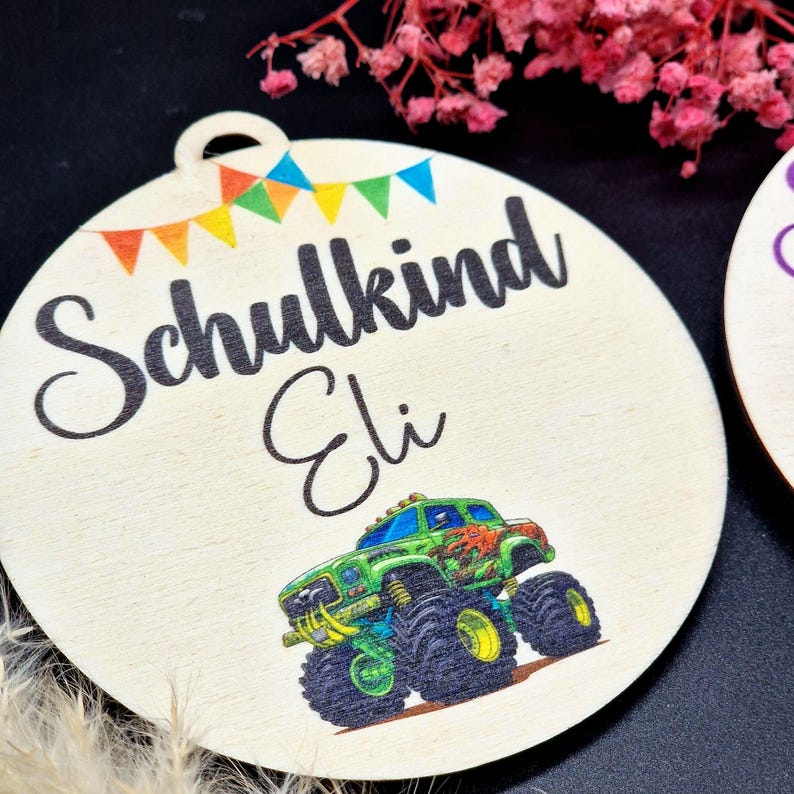 Pode incluir: Enfeite redondo de madeira com o texto "Schulkind Eli" e uma ilustra&ccedil;&atilde;o de um monster truck colorido. O enfeite tem uma argola no topo e uma faixa de bandeiras coloridas.