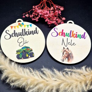 Pode incluir: Dois enfeites redondos de madeira com um furo no topo. Um apresenta um desenho de monster truck e o nome "Eli" com a palavra "Schulkind". O outro mostra um cavalo e o nome "Nele" com a palavra "Schulkind".