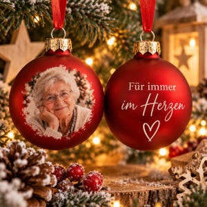 Pode incluir: Duas esferas de Natal vermelhas foscas com tampas douradas e fitas vermelhas. Uma esfera apresenta uma foto circular de uma pessoa idosa de óculos, enquanto a outra exibe o texto branco "Für immer im Herzen" acima de um coração desenhado.