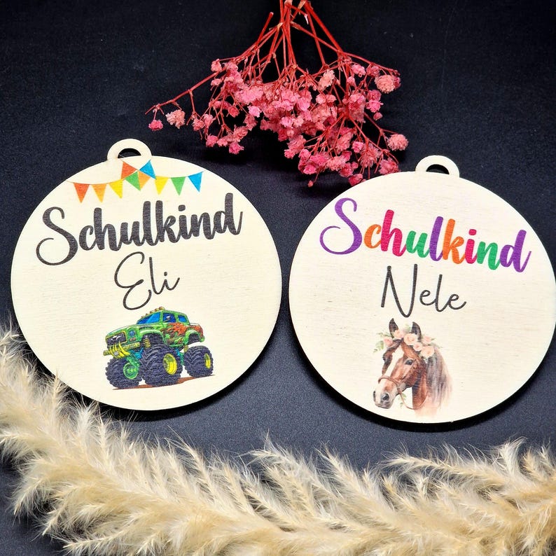 Pode incluir: Dois enfeites redondos de madeira com a palavra alem&atilde; "Schulkind" e os nomes "Eli" e "Nele". Um apresenta um monster truck, o outro um cavalo com uma coroa floral. Ambos t&ecirc;m um buraco para pendurar.