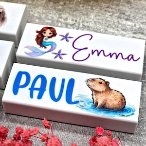 May include: White rectangular blocks with custom designs. One block features a mermaid illustration with the name "Emma" in purple script. Another block displays the name "PAUL" in blue, with a capybara illustration.