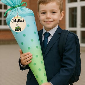 Pode incluir: Uma grande mochila escolar em forma de cone em tons de azul e verde, decorada com bolinhas verdes. A mochila &eacute; amarrada com uma fita azul-petr&oacute;leo e uma etiqueta de madeira que diz "Schulkind Eli" com uma ilustra&ccedil;&atilde;o de um monster truck. Uma crian&ccedil;a de terno azul-marinho segura a mochila.