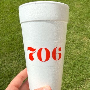 May include: A white styrofoam cup featuring the number 706 printed in a bold red font on the front. The cup has a textured surface and a tapered cylindrical shape with a wide rim at the top, designed for holding cold beverages.