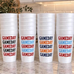 May include: Four stacks of white foam cups, each featuring the word GAMEDAY printed five times in various colors. The first stack has black and red text, the second has blue and red, the third has navy and orange, and the fourth has blue and red.