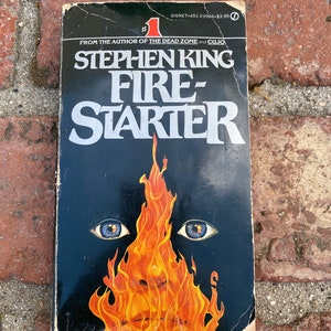 May include: A paperback book titled "Firestarter" by Stephen King. The cover features a black background with a fiery face and blue eyes. The book is a Signet edition.