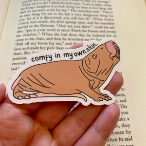 May include: A sticker of a brown naked mole rat with the text "comfy in my own skin." The mole rat is lying on its belly with its mouth open.
