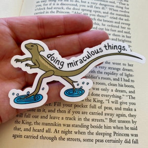 May include: A sticker featuring a cartoon lizard walking on water with the text "doing miraculous things." The lizard is brown and green, and the water is blue. The sticker is on a book.