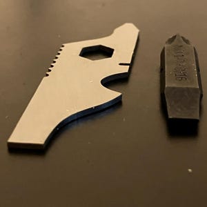 以下が含まれることがあります： 暗い表面に、シルバーのマルチツールと黒いドライバービットが写っています。マルチツールは六角形の穴、鋸のようなエッジ、栓抜きを備えています。ドライバービットには「N112-8/16」の文字が刻まれています。