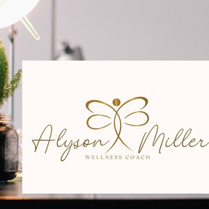K&ouml;nnte beinhalten: Goldenes Logo auf cremefarbener Karte. Der Name "Alyson Miller" ist in Schreibschrift geschrieben, darunter steht "Wellness Coach". Das Logo zeigt eine stilisierte Libellenfigur.