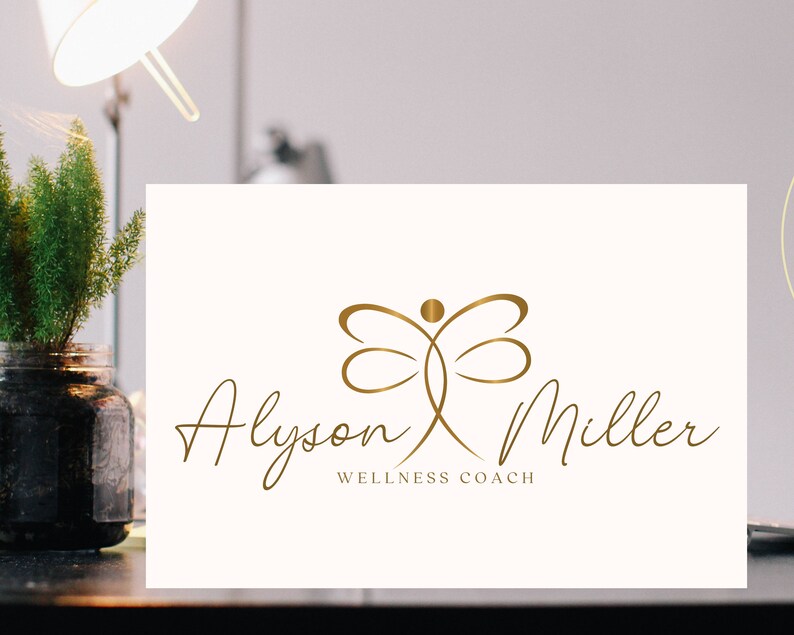 K&ouml;nnte beinhalten: Goldenes Logo auf cremefarbener Karte. Der Name "Alyson Miller" ist in Schreibschrift geschrieben, darunter steht "Wellness Coach". Das Logo zeigt eine stilisierte Libellenfigur.