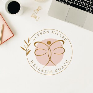 K&ouml;nnte beinhalten: Draufsicht auf einen Arbeitsplatz mit Laptop, Kaffeetasse, rosa Notizbuch und goldenem Stift. Das Logo von Alyson Miller Wellness Coach mit einem goldenen Schmetterlingsdesign in einem rosa Kreis ist deutlich sichtbar.