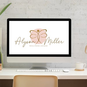 K&ouml;nnte beinhalten: Ein Computermonitor zeigt das Logo "Alyson Miller Wellness Coach". Das Logo zeigt ein goldenes Schmetterlingsdesign &uuml;ber einem rosa Kreis mit goldenem Schriftzug. Der Monitor steht auf einem wei&szlig;en Schreibtisch mit einer Pflanze, einem Telefon und einer Kaffeetasse.