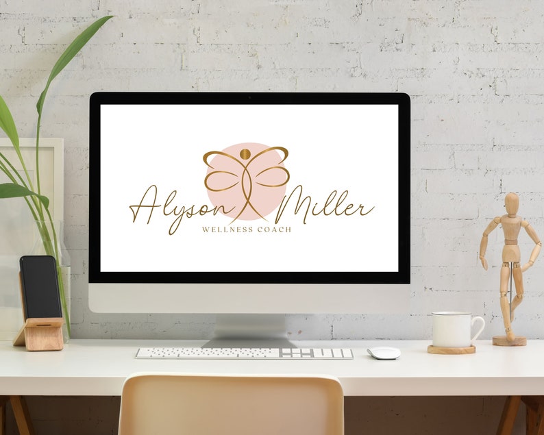 K&ouml;nnte beinhalten: Ein Computermonitor zeigt das Logo "Alyson Miller Wellness Coach". Das Logo zeigt ein goldenes Schmetterlingsdesign &uuml;ber einem rosa Kreis mit goldenem Schriftzug. Der Monitor steht auf einem wei&szlig;en Schreibtisch mit einer Pflanze, einem Telefon und einer Kaffeetasse.