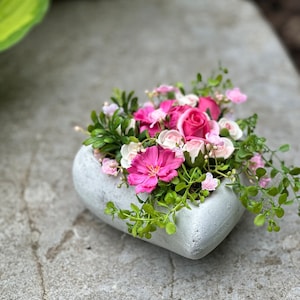 Könnte beinhalten: Ein herzförmiger Betontopf gefüllt mit rosa und weißen Blumen, darunter Rosen und andere Blüten. Der Topf steht auf einer grauen Steinoberfläche.