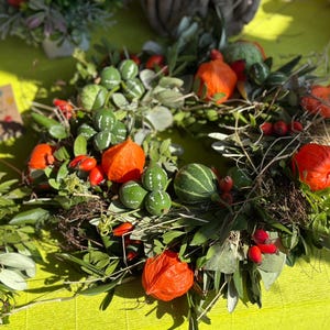 May include: A circular wreath constructed from green foliage, featuring small striped green gourds, bright orange physalis husks, and clusters of red berries. The arrangement is woven with thin brown twigs and stems, creating a textured, natural appearance.