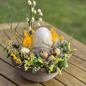 Könnte beinhalten: Ein dekoratives Ostergesteck in einer braunen Schale. Es zeigt ein großes, perlmuttweißes Ei, gefleckte Wachteleier, gelbe Blüten und Zweige mit Kätzchen. Die Anordnung befindet sich auf einem Holztisch mit verschwommenem grünem Hintergrund.
