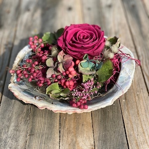 Könnte beinhalten: Ein Blumengesteck in einer großen, schillernden Muschelschale. Es besteht aus einer tiefrosa Rose, kleinen roten Beeren und grünen Blättern. Die Schale liegt auf einer verwitterten Holzoberfläche. Durchmesser ca. 20 cm.