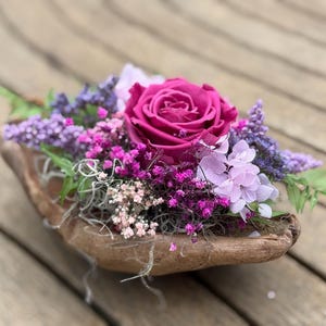 Może przedstawiać: Drewniana miska wypełniona żywą kompozycją kwiatową. Kompozycja zawiera dużą, głęboką różową różę jako punkt centralny, otoczoną mniejszymi fioletowymi, różowymi i białymi kwiatami oraz zielonymi liśćmi. Miska stoi na drewnianej powierzchni.