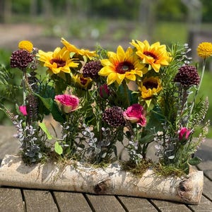 Pode incluir: Arranjo floral com girassóis, rosas cor-de-rosa e flores bordô em um recipiente de casca de bétula. O arranjo inclui vegetação e está sobre uma mesa de madeira, criando uma estética natural.