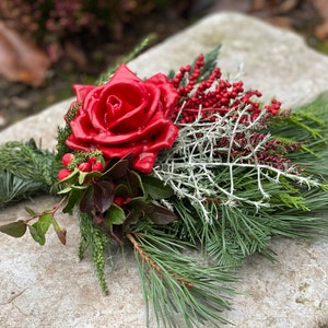 Könnte beinhalten: Ein handgefertigtes Blumenarrangement mit einer großen, glänzenden roten Rose als Mittelpunkt. Das Arrangement umfasst grüne Tannennadeln, rote Beeren und silbergraues Laub, wodurch eine natürliche und festliche Ästhetik entsteht. Der Strauß ist mit grünem Bindfaden gebunden.