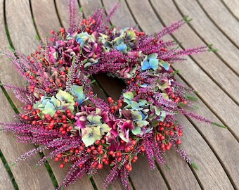 May include: A circular wreath composed of vibrant purple heather sprigs, clusters of small red berries, and dried hydrangea blossoms in shades of deep magenta, muted blue, and soft green, arranged in a dense, textured, and multi-layered floral display.