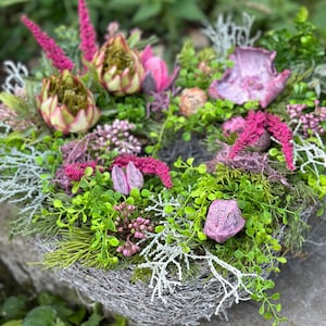 Può includere: Una ghirlanda a forma di cuore realizzata con fiori secchi e verde. La ghirlanda è decorata con fiori rosa e viola, oltre a foglie verdi. La ghirlanda è appoggiata su una roccia grigia.