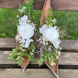 Può includere: Due composizioni floreali decorative con rose bianche, fogliame verde ed elementi secchi. I fiori sono disposti in uno stile naturale e rustico, su uno sfondo di legno. Ideale per l'arredamento della casa o eventi speciali.