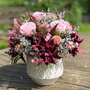 Könnte beinhalten: Ein Blumenarrangement in einer weißen, strukturierten Vase. Der Strauß zeigt rosa Rosen, burgunderfarbene Dahlien und verschiedene andere Blumen in Rosa-, Burgunder- und Grüntönen. Das Arrangement steht auf einem Holztisch.