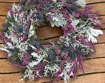 May include: A circular wreath composed of vibrant magenta heather sprigs, spiky blue-gray sea holly thistle heads, and silvery-white dusty miller foliage. The arrangement features a dense, textured mix of these botanical elements.