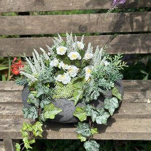Könnte beinhalten: Eine dunkelgraue, steinartige Schale mit einem Arrangement aus weißen Blüten, grünen Efeuranken und hohen, schlanken weißen Blütenähren. Die Efeublätter weisen hell- und dunkelgrüne Muster auf und hängen über die Seiten der Schale herab.