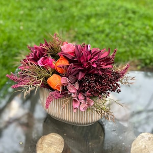 Könnte beinhalten: Ein herbstliches Blumenarrangement in einer gestreiften Keramikvase. Das Arrangement besteht aus burgunderfarbenen Dahlien, rosa Rosen, orangefarbenen mohnartigen Blüten und burgunderfarbenen Beeren.