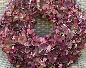 May include: A circular wreath constructed from dried hydrangea blossoms in deep shades of burgundy, plum, and muted green. The dense arrangement features numerous small, overlapping petals creating a full, textured, and rounded floral display.
