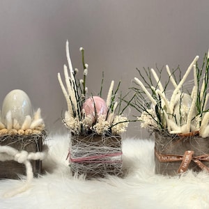 Könnte beinhalten: Drei dekorative Osterarrangements in kleinen Holzkisten. Jede Kiste enthält ein Ei, umgeben von getrockneten Blumen und Zweigen. Die Kisten sind mit Bindfaden und Schleifen gebunden und stehen auf einer weißen, flauschigen Oberfläche. Die Arrangements sind in neutralen Tönen gehalten.