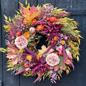 Può includere: Una ghirlanda autunnale con una varietà di fiori secchi, foglie ed erbe. La ghirlanda è decorata con rose rosa, dalie bordeaux e bacche arancioni.